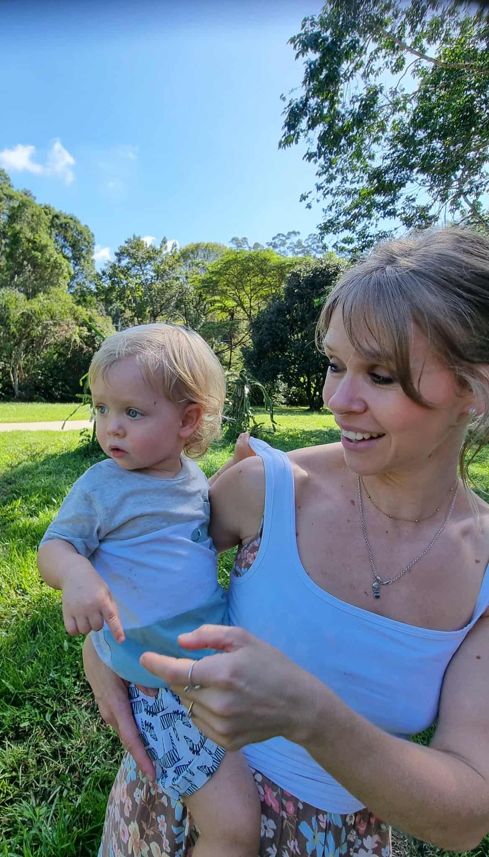 Sally smiling with her youngest son on her hip in a sunny green park